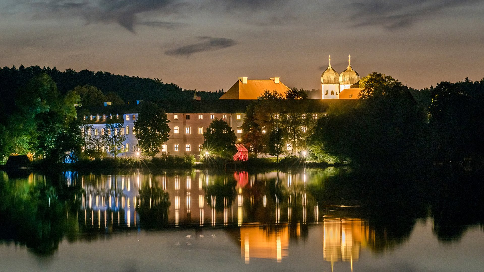 Kloster Seeon bei Nacht mit beleuchteten Gebäuden und Kirchtürmen, Spiegelung im See, umgeben von Bäumen und dunklem Himmel.