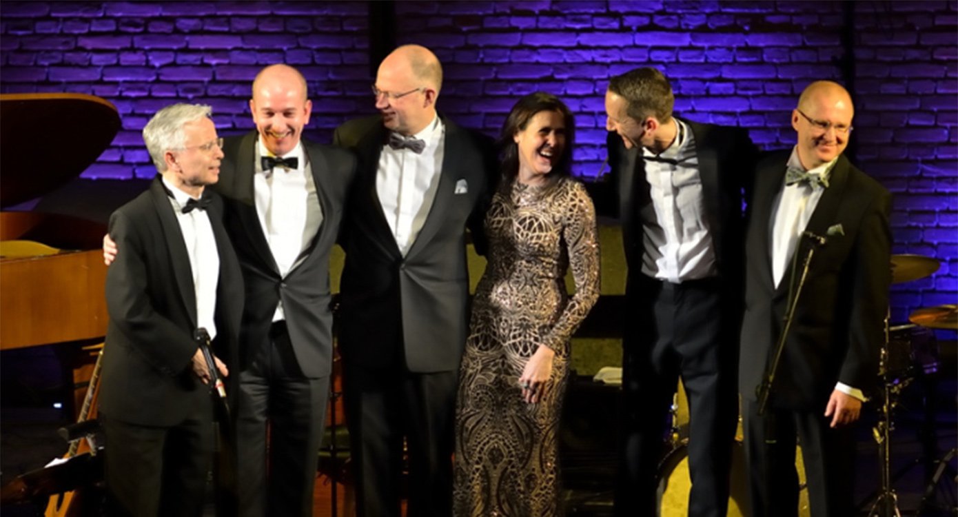 Fünf Herren und eine Dame in Abendkleid auf einer Bühne vir den Instrumenten stehend