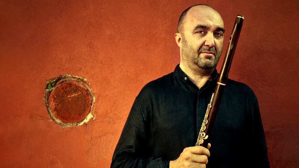 Foto einer Person in schwarzem Hemd vor rostrot strukturierter Wand, die eine Querflöte senkrecht in den Händen hält, daneben ein rundes Metallelement in der Wand zur Veranstaltung Mozart Matinee Mercelli Massimo
