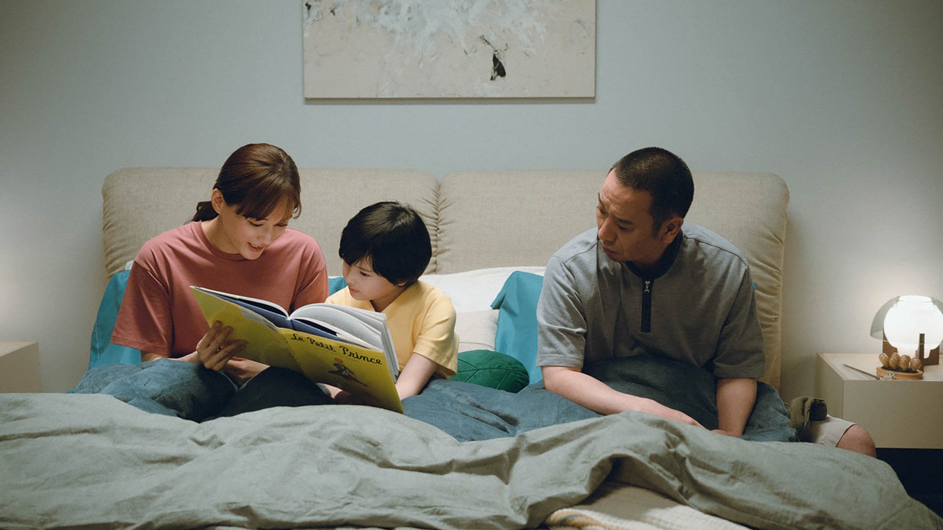 Drei Personen sitzen gemeinsam auf einem Bett; eine Person in der Mitte liest aus einem aufgeschlagenen Buch, während die beiden anderen seitlich daneben sitzen und zuhören; im Hintergrund sind eine helle Wand und ein Bild zu sehen.
