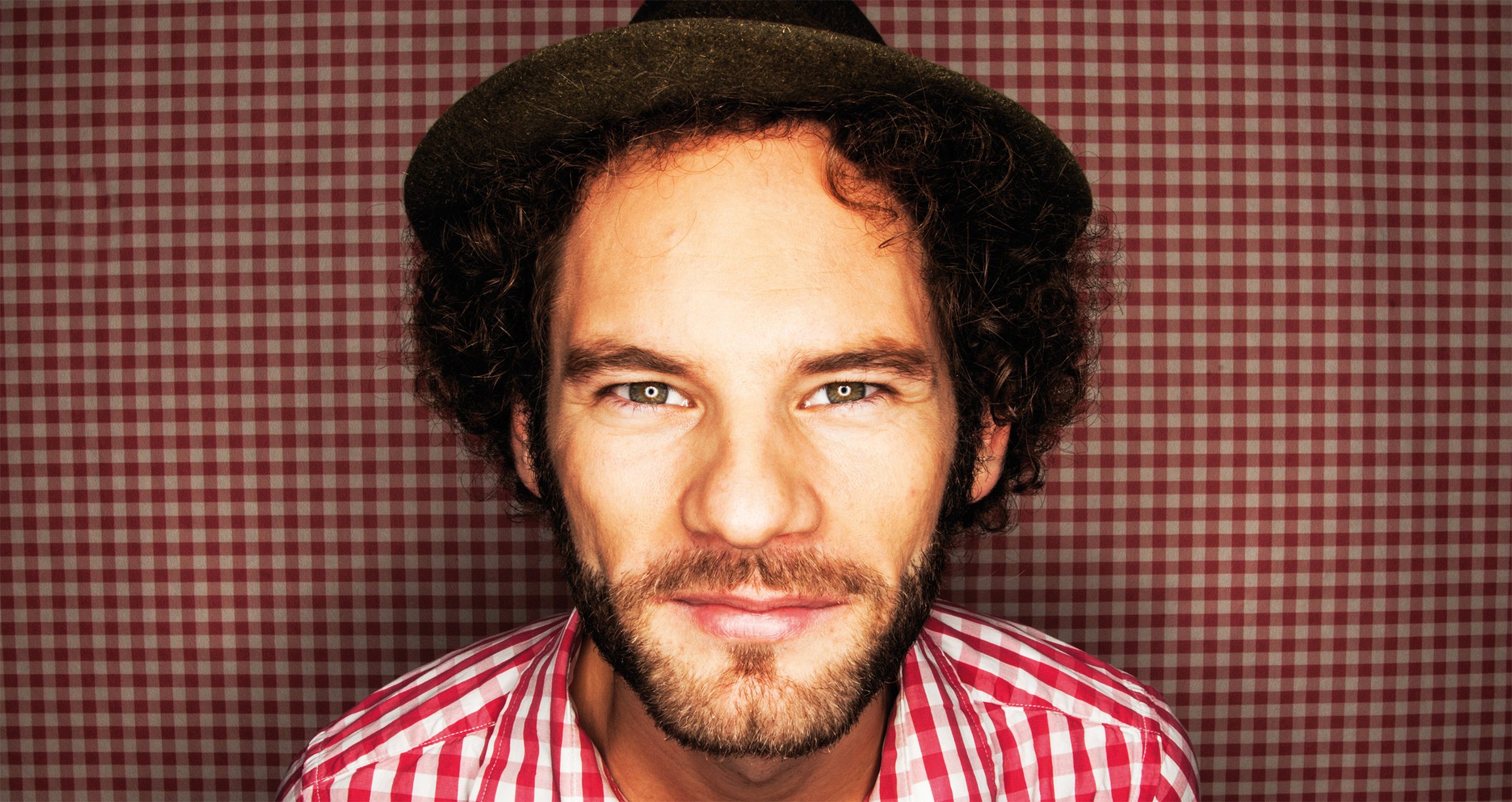 Foto für die Veranstaltung von Maxi Schafroth – Faszination Bayern mit dem Künstler in einem rot‑weiß karierten Hemd und demselben karierten Hintergrund