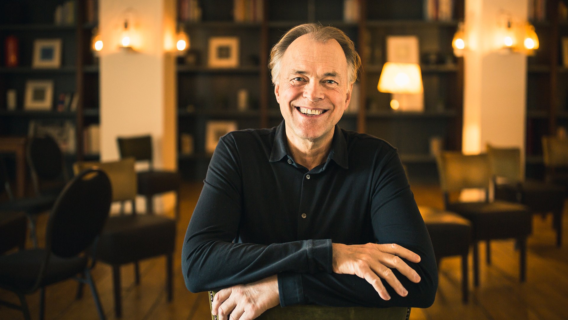 Der Dirigent Thomas Hengelbrock sitzt an einem Tisch in einem warm beleuchteten Raum mit Stühlen und Bücherregalen im Hintergrund.