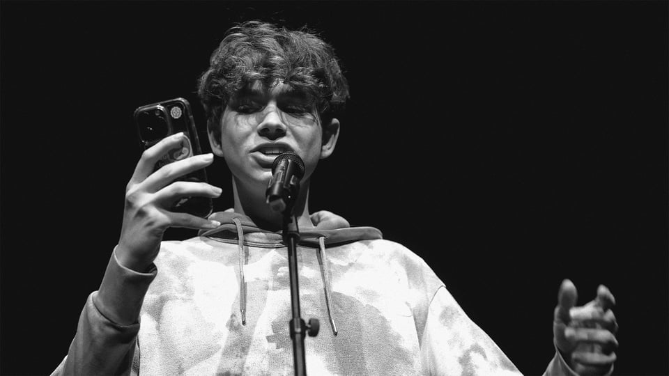 Foto für das Event „U20 POETRY SLAM“ in Schwarz-Weiß, mit einem Jungen, der mit Mikrofon und Handy performt.