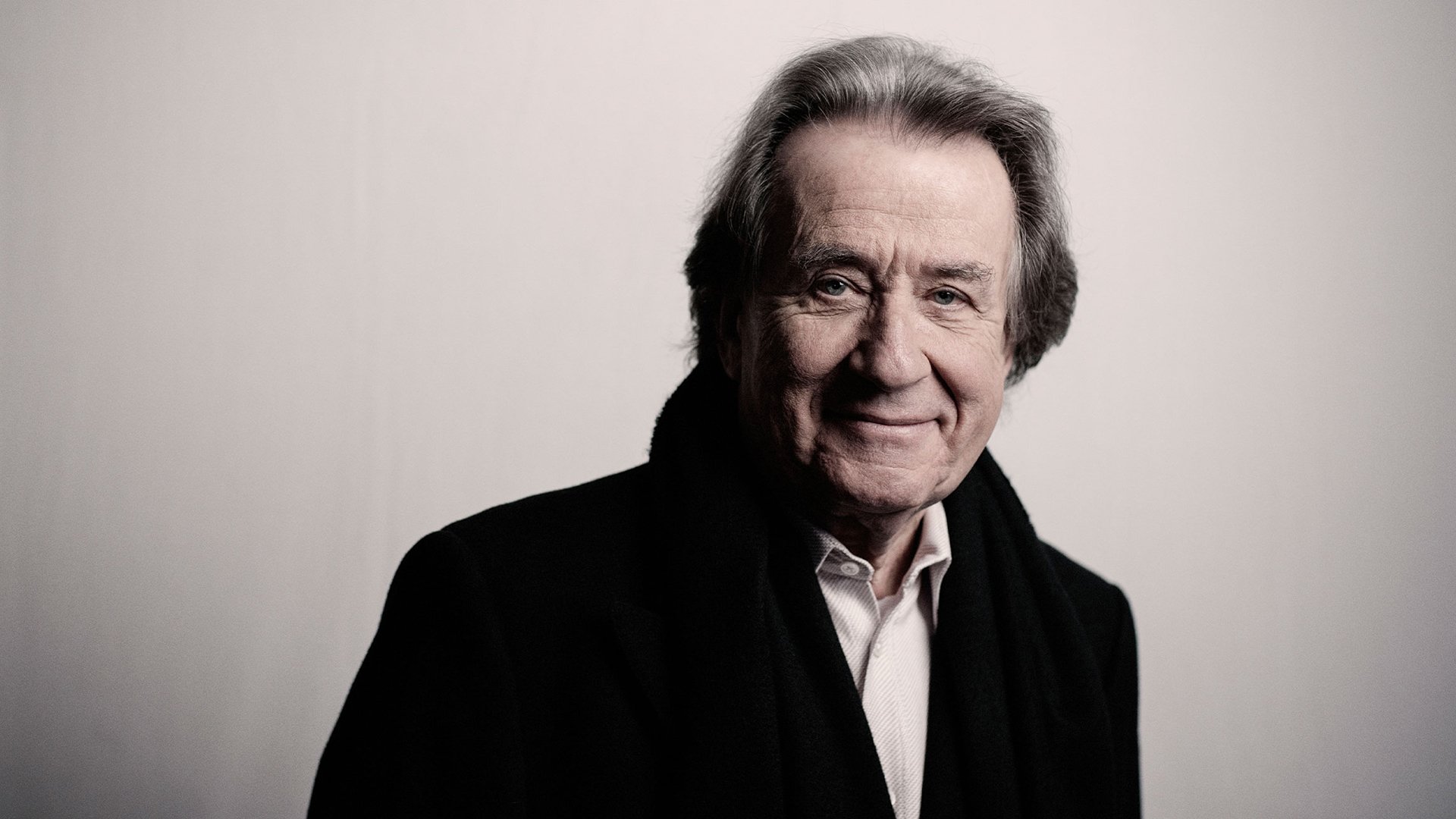 Portrait von Rudolf Buchbinder, lächelnd in hellem Hemd und schwarzem Mantel vor hellgrauem Hintergrund
