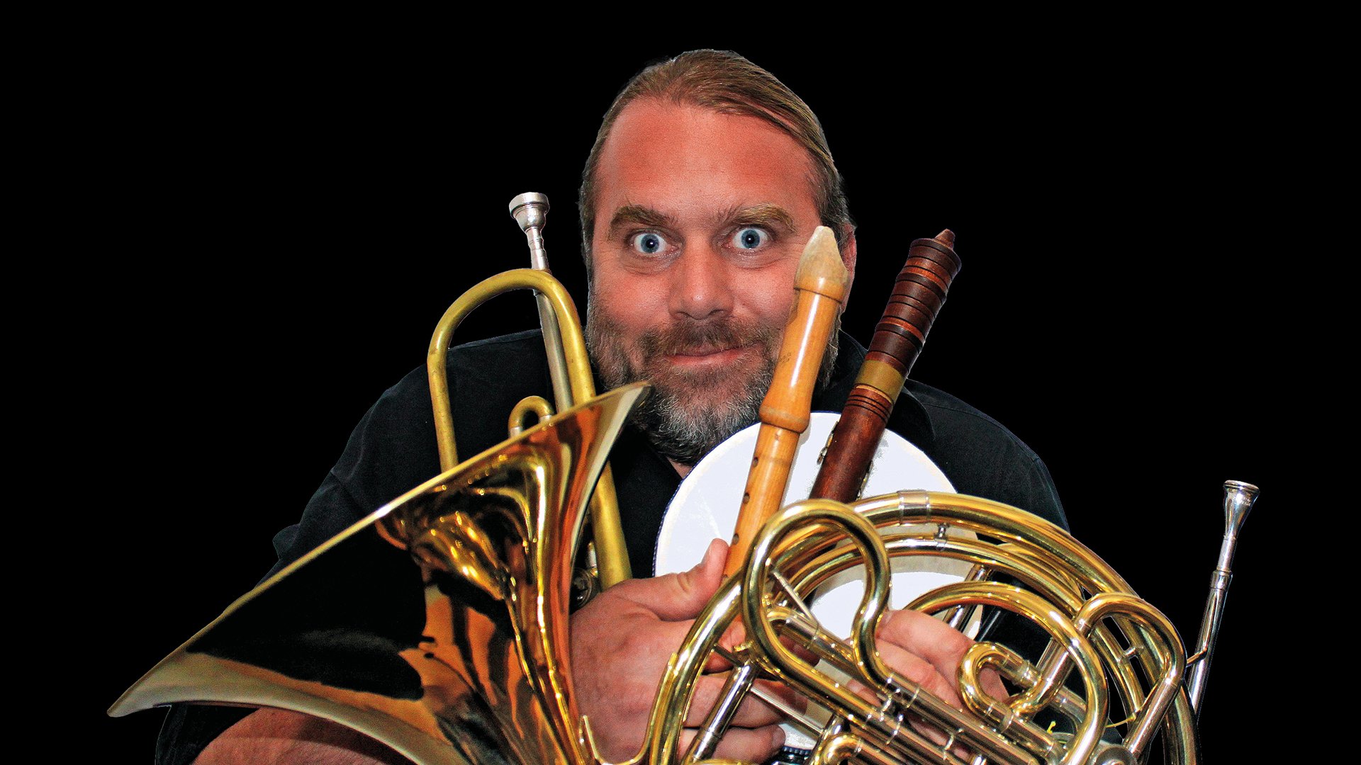 Andreas Martin Hofmeir mit verschiedenen Blasinstrumenten schneidet eine Grimasse in die Kamera, vor schwarzem Hintergrund