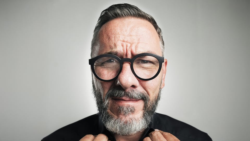 Nahaufnahme einer Person mit Brille und Bart vor neutralem Hintergrund, Porträtfoto für ein Comedy‑Programm.
