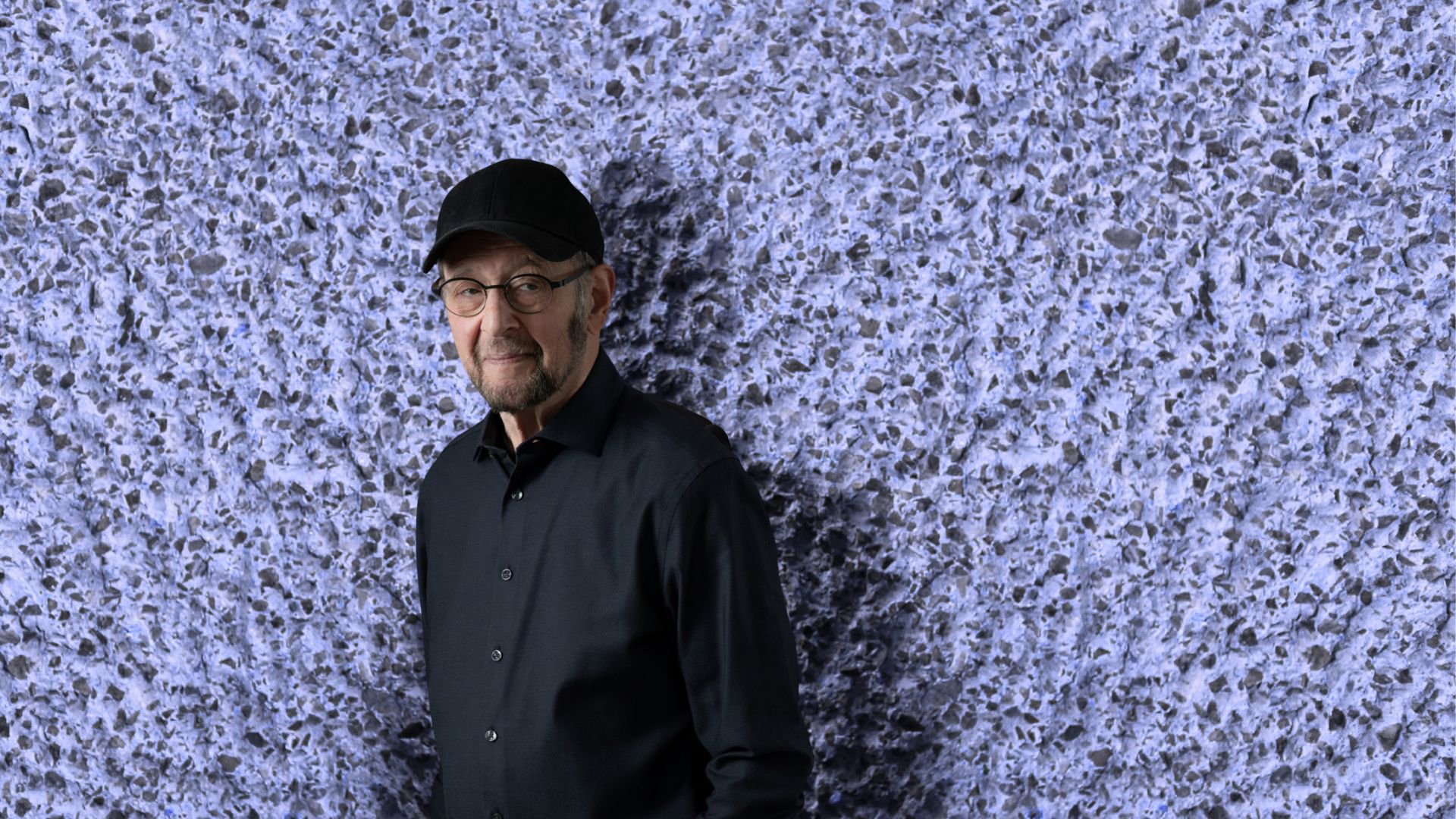 Eine Person in schwarzem Hemd und schwarzer Kappe vor violetter rauher Steinwand zur Veranstaltung KOMPONISTENPORTRÄT STEVE REICH