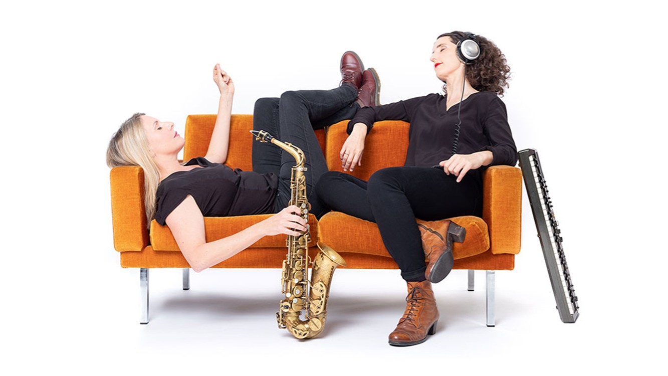 Pressefoto des Duos Breuer & Hermenau mit zwei Musikerinnen vor weißem Hintergrund auf einem orangefarbenen Sofa, eine mit Saxofon liegend auf dem Sofa, die andere mit Kopfhörern sitzend, daneben ein Akkordeon.