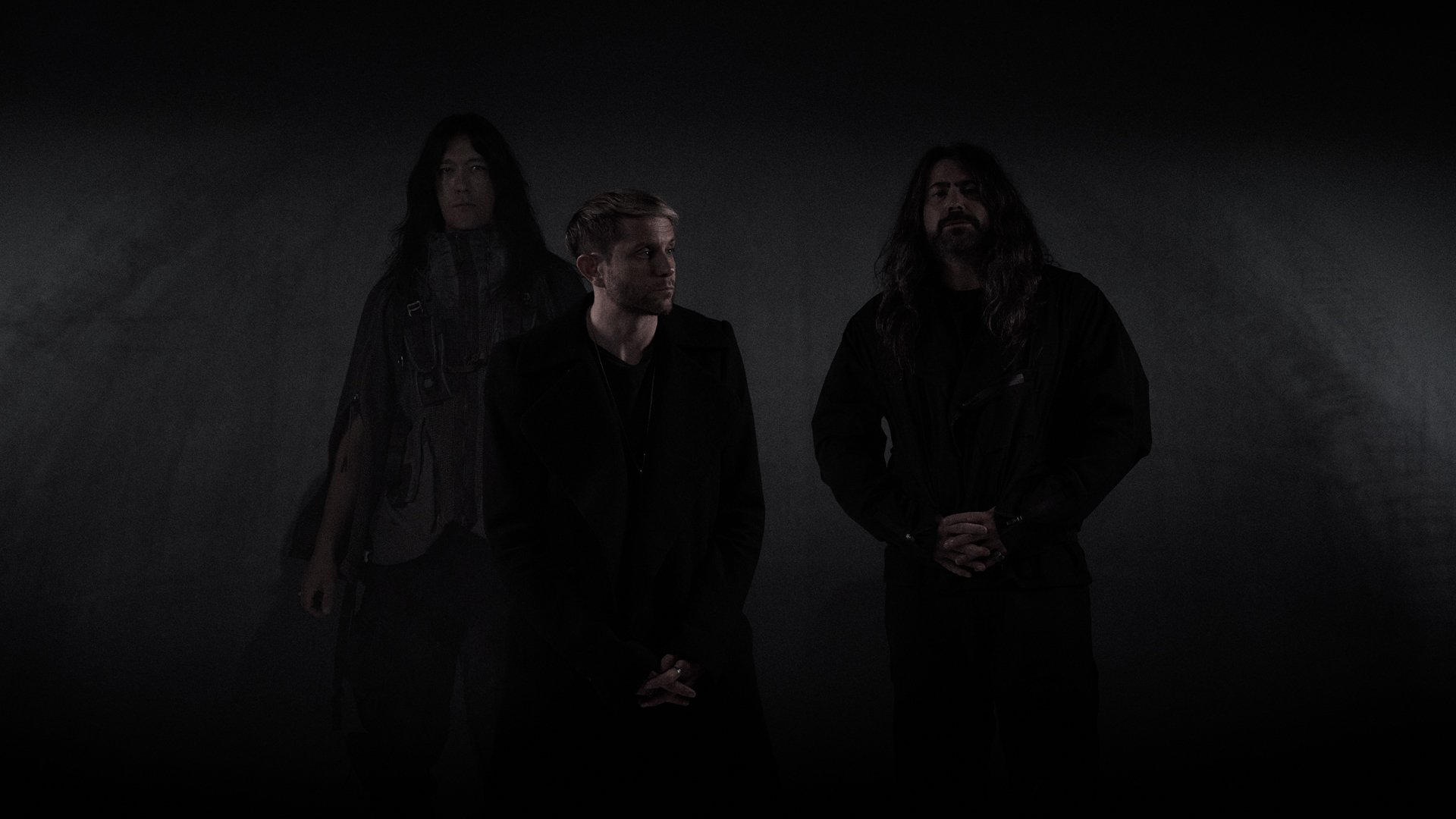 Drei Mitglieder der Band HEALTH stehen vor einem dunklen Hintergrund und blicken ernst; sie tragen schwarze Kleidung und sind minimalistisch beleuchtet, was eine düstere, atmosphärische Stimmung erzeugt.