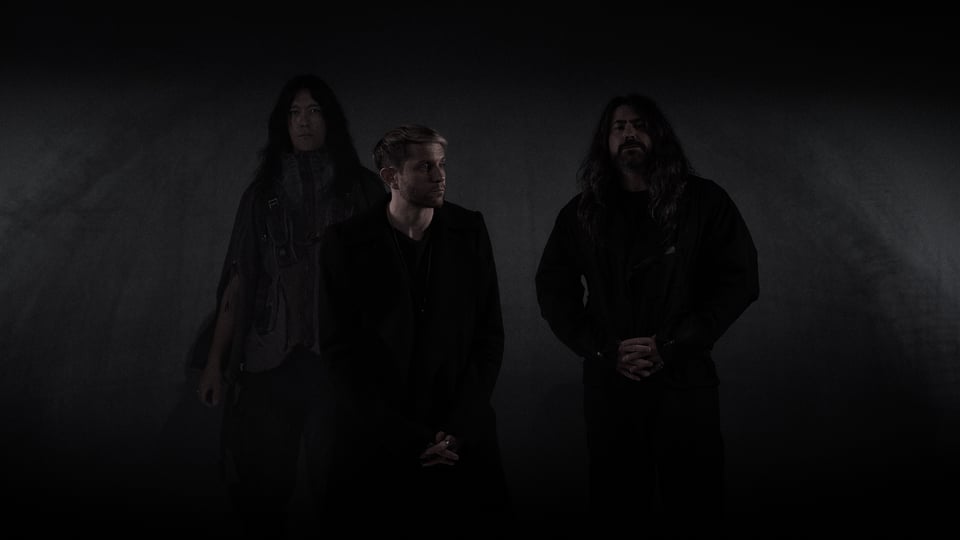 Drei Mitglieder der Band HEALTH stehen vor einem dunklen Hintergrund und blicken ernst; sie tragen schwarze Kleidung und sind minimalistisch beleuchtet, was eine düstere, atmosphärische Stimmung erzeugt.