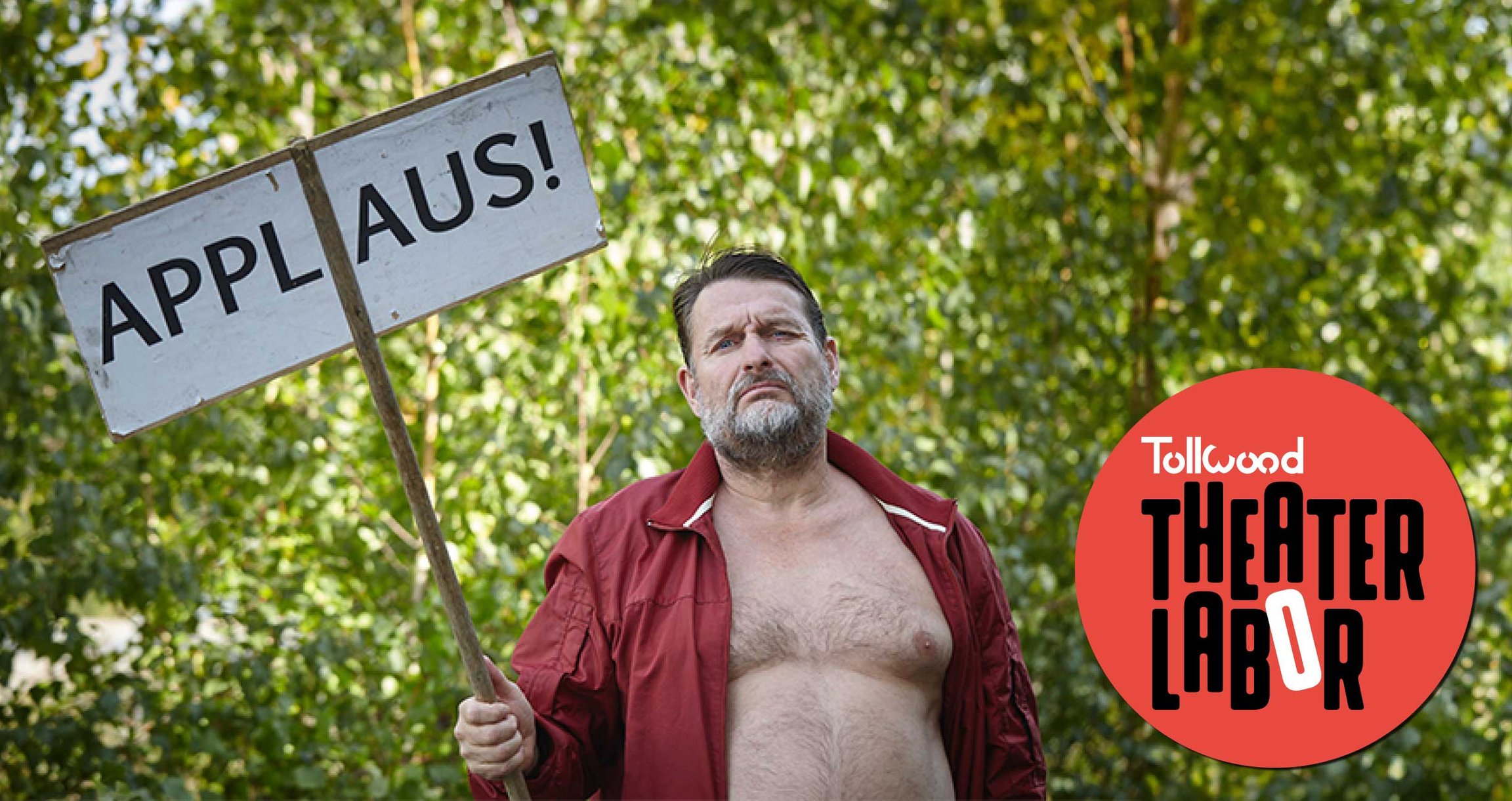 Mann mit rotem Hemd und Applaus-Holzschild vor Gebüsch