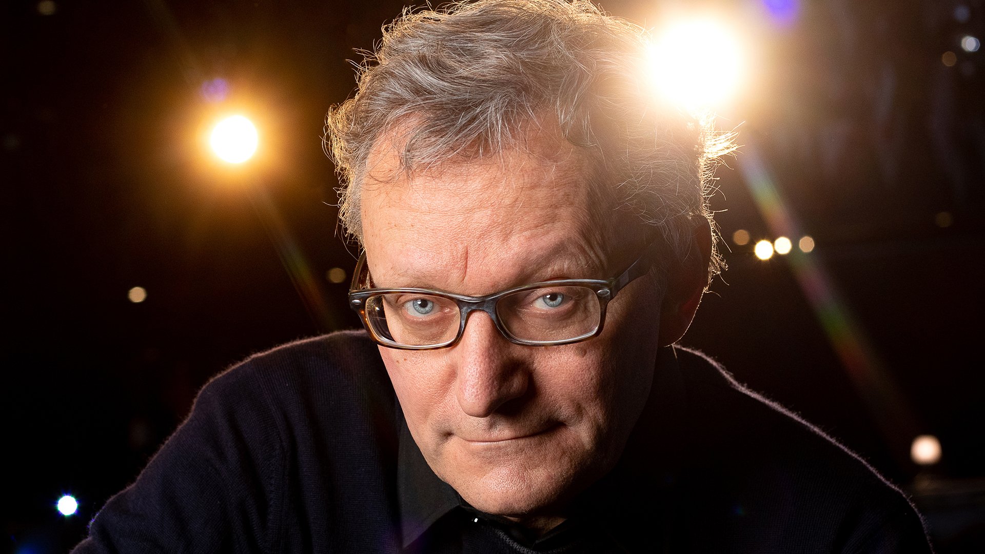 Porträtfoto von Frank Lüdecke in dunklem Pullover und Brille vor unscharfem Bühnenhintergrund, mit direktem Blick zur Kamera und hellen Scheinwerfern im Hintergrund