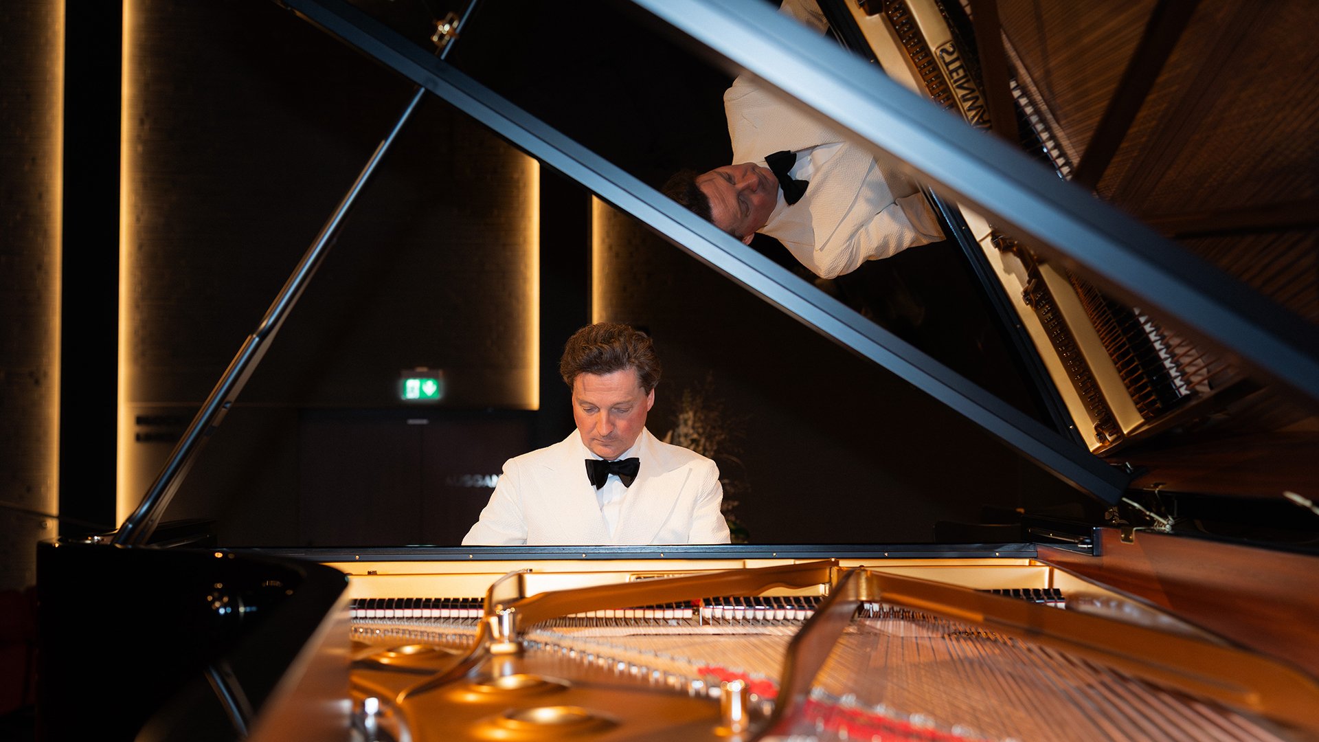 Ein Pianist im weißen Smoking mit schwarzer Fliege an einem geöffneten Konzertflügel, aufgenommen durch den Flügelrahmen mit Spiegelung im aufgeklappten Deckel, vor warm beleuchtetem Konzertsaalhintergrund.