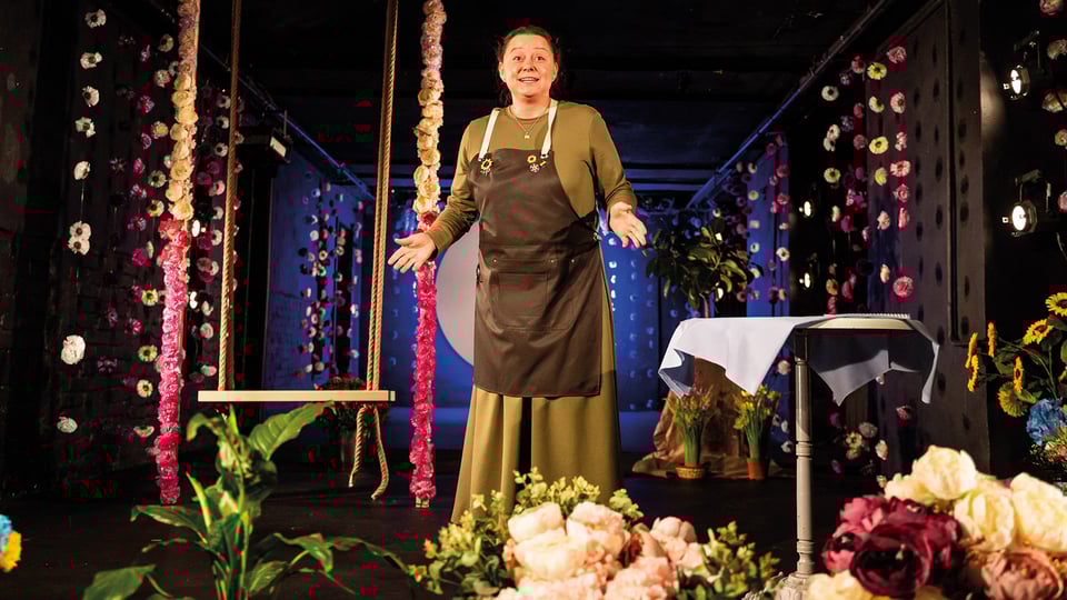 Foto für die Veranstaltung „Soloway 2026 – Love Me Don’t Leave“ mit einer Darstellerin zwischen Blumen und einem Tisch neben ihr.