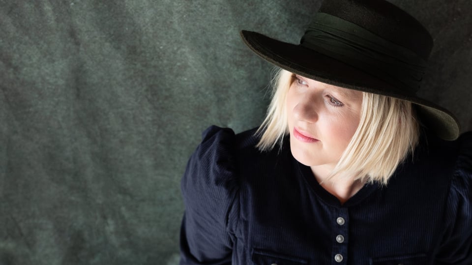 Claudia Koreck mit schwarzem Hut und dunkelblauem Oberteil blickt zur Seite, vor grau marmoriertem Hintergrund