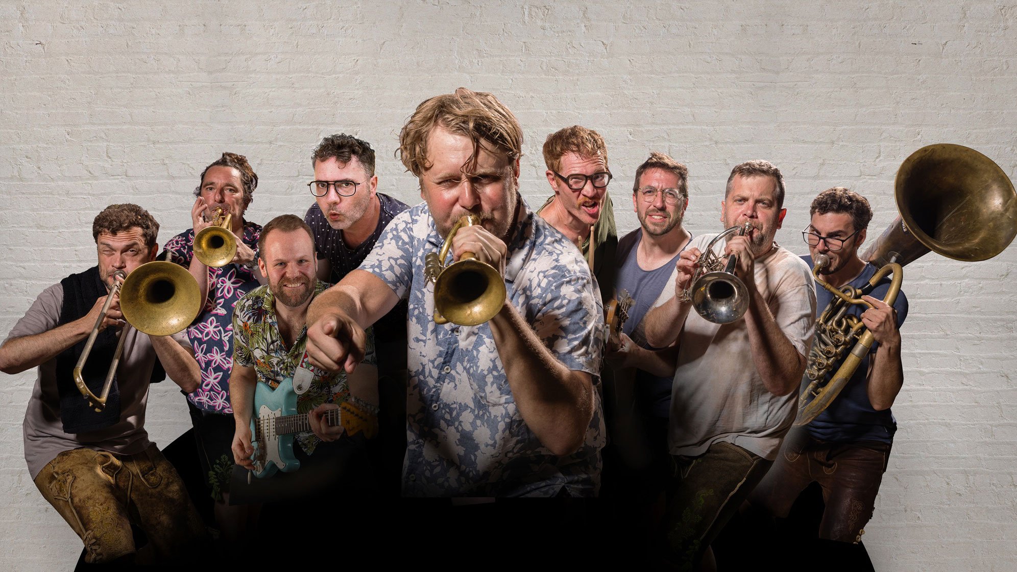Mitglieder von LaBrassBanda mit Trompeten, Posaune, Tuba und E Gitarre vor einer weißen Backsteinwand.