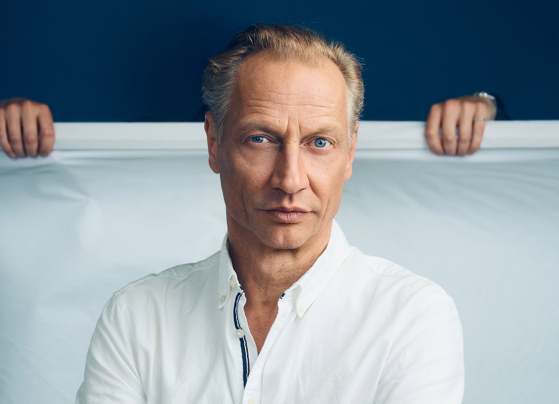 Foto eines Mannes mit weißem Hemd und im Hintergrund eine Hand, die ein weißes Tuch hält, das ihn bedeckt.