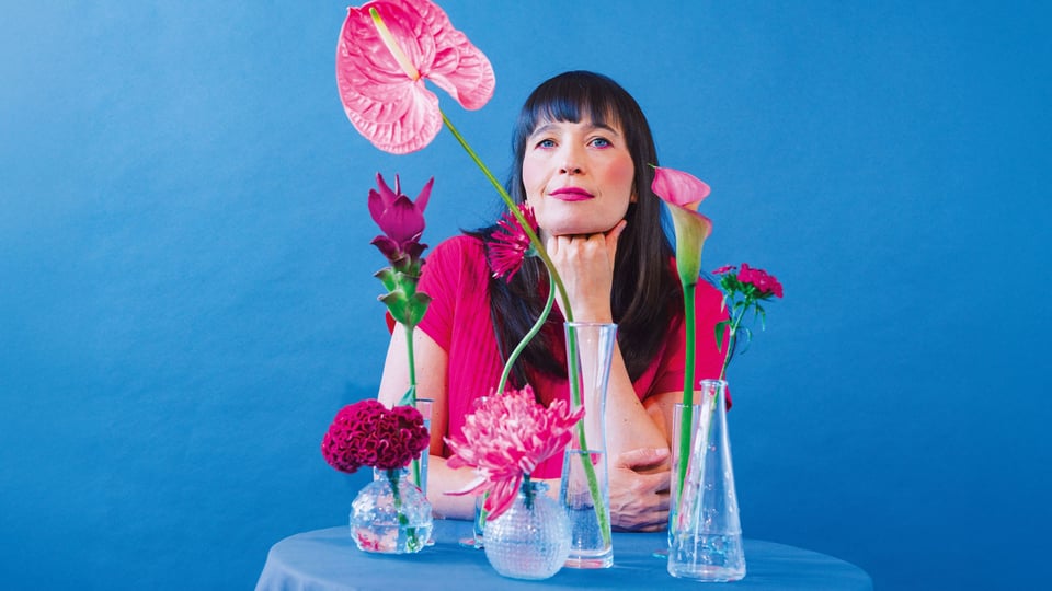 Die Künstlerin Hannah Köpf in pinkem Pullover sitzt an einem Tisch mit blauer Tischdecke und Glasvasen mit verschiedenen pinken Blumen, vor blauem Hintergrund