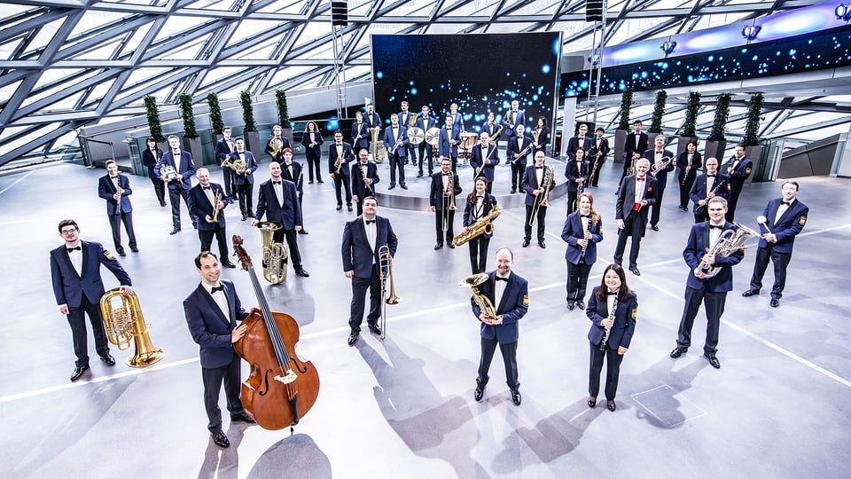 Große Musikergruppe mit verschiedenen Blas‑, Schlag‑ und Streichinstrumenten steht in formierter Aufstellung in einem modernen, lichtdurchfluteten Saal mit Glasdach.