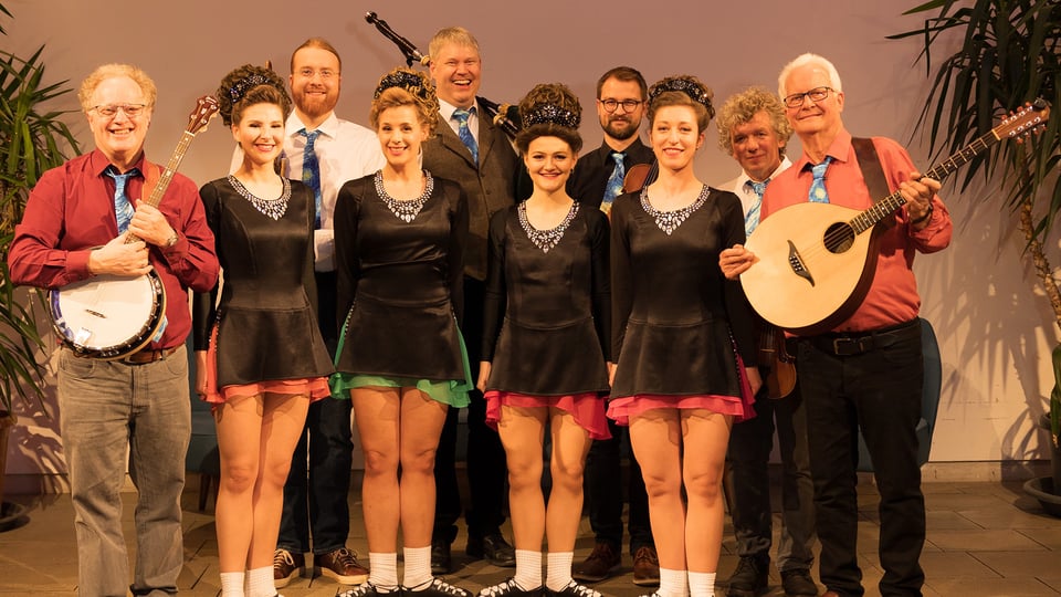 Musikensemble mit Instrumentalisten und Tänzern steht in Reihe auf Bühne, einige halten Banjo und Mandoline, mehrere tragen koordinierte Bühnenkostüme.