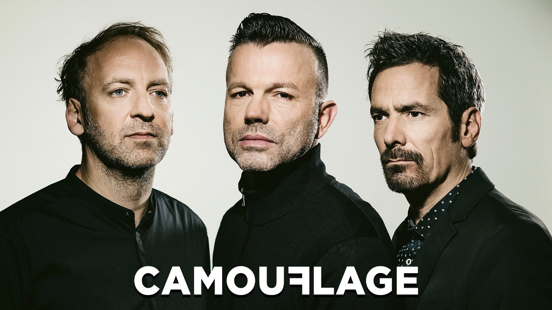 Eventmotiv der Band Camouflage: Drei Personen stehen dicht nebeneinander vor hellem, neutralem Hintergrund und blicken frontal in die Kamera. Dunkle, schlichte Kleidung mit klaren Linien. Unten im Bild ist der weiße Schriftzug „CAMOUFLAGE“ zu sehen. Ruhiges, reduziertes Porträtmotiv für eine Live‑Konzertankündigung.