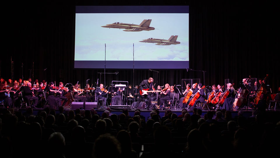 Bühnenfoto eines Orchesters mit Streichern und Gitarrist auf einer Konzertbühne vor Publikum, darüber eine große Leinwand mit der Projektion zweier Kampfjets über den Wolken zur Veranstaltung The Magic of Hans Zimmer