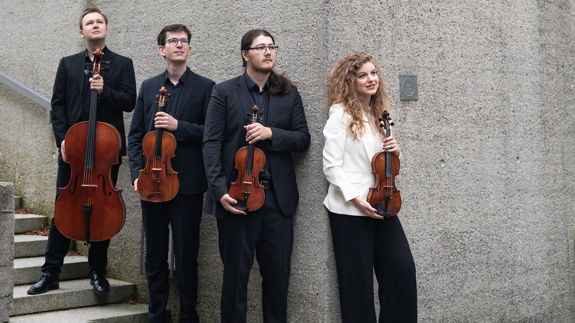 Gruppenfoto eines Streichquartetts im Außenbereich: Vier Personen stehen nebeneinander vor einer grauen Betonwand und halten klassische Streichinstrumente – links ein Violoncello, daneben zwei Violinen, rechts eine Viola. Die Personen tragen überwiegend dunkle, elegante Kleidung, eine Person rechts trägt eine helle Jacke. Die Aufnahme ist ruhig komponiert, mit natürlichem Licht und klarer Fokussierung auf Instrumente, Haltung und Ensemble‑Charakter, geeignet als Künstler‑ oder Konzertfoto.