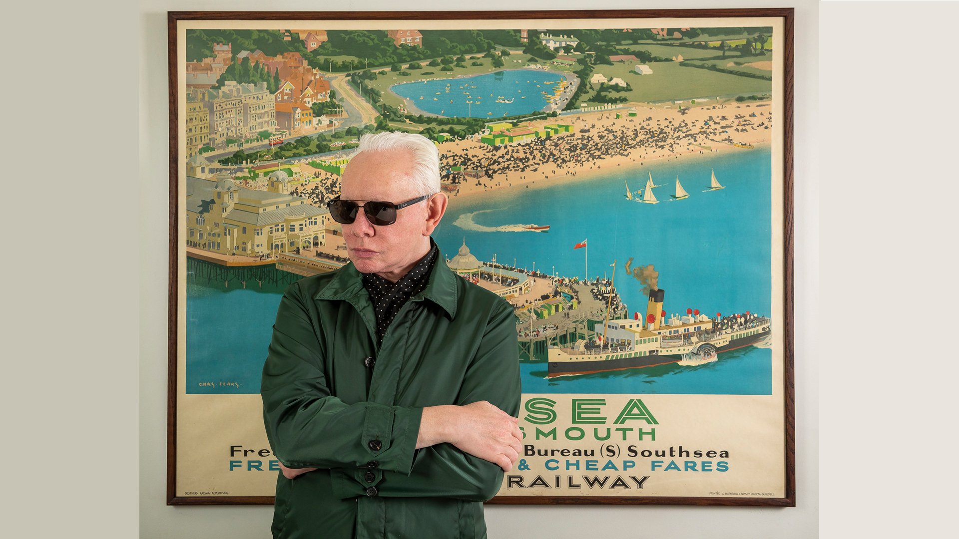 Der Künstler steht mit Sonnenbrille vor einem nostalgischen Plakat einer Bucht mit Hafen