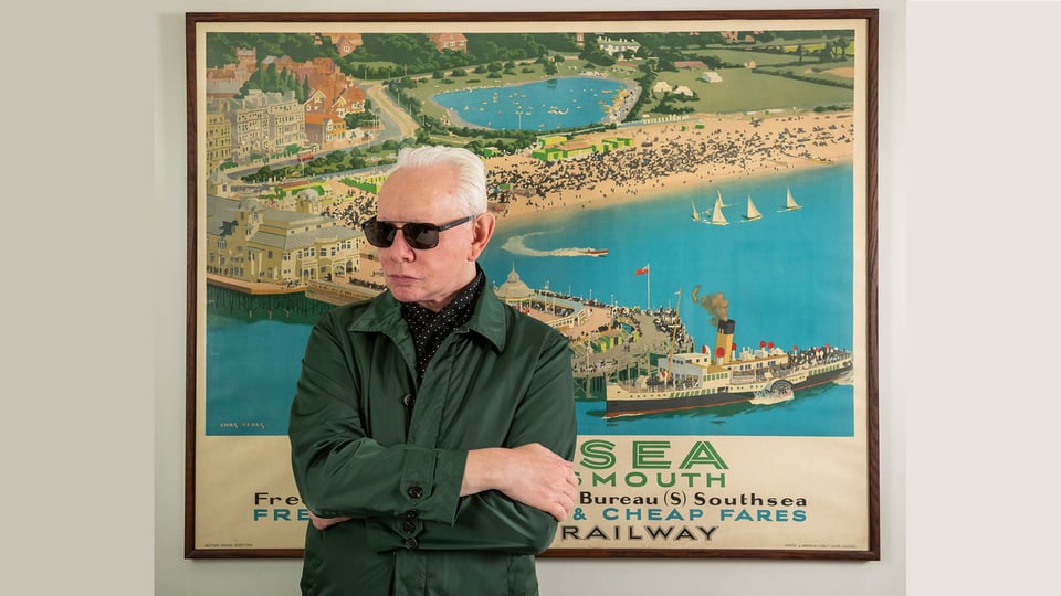 Der Künstler steht mit Sonnenbrille vor einem nostalgischen Plakat einer Bucht mit Hafen