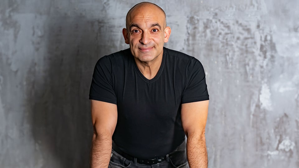 Der Kabarettist Django Asyl in schwarzem Shirt nach vorne gestützt vor grauem verschwommenem Hintergrund