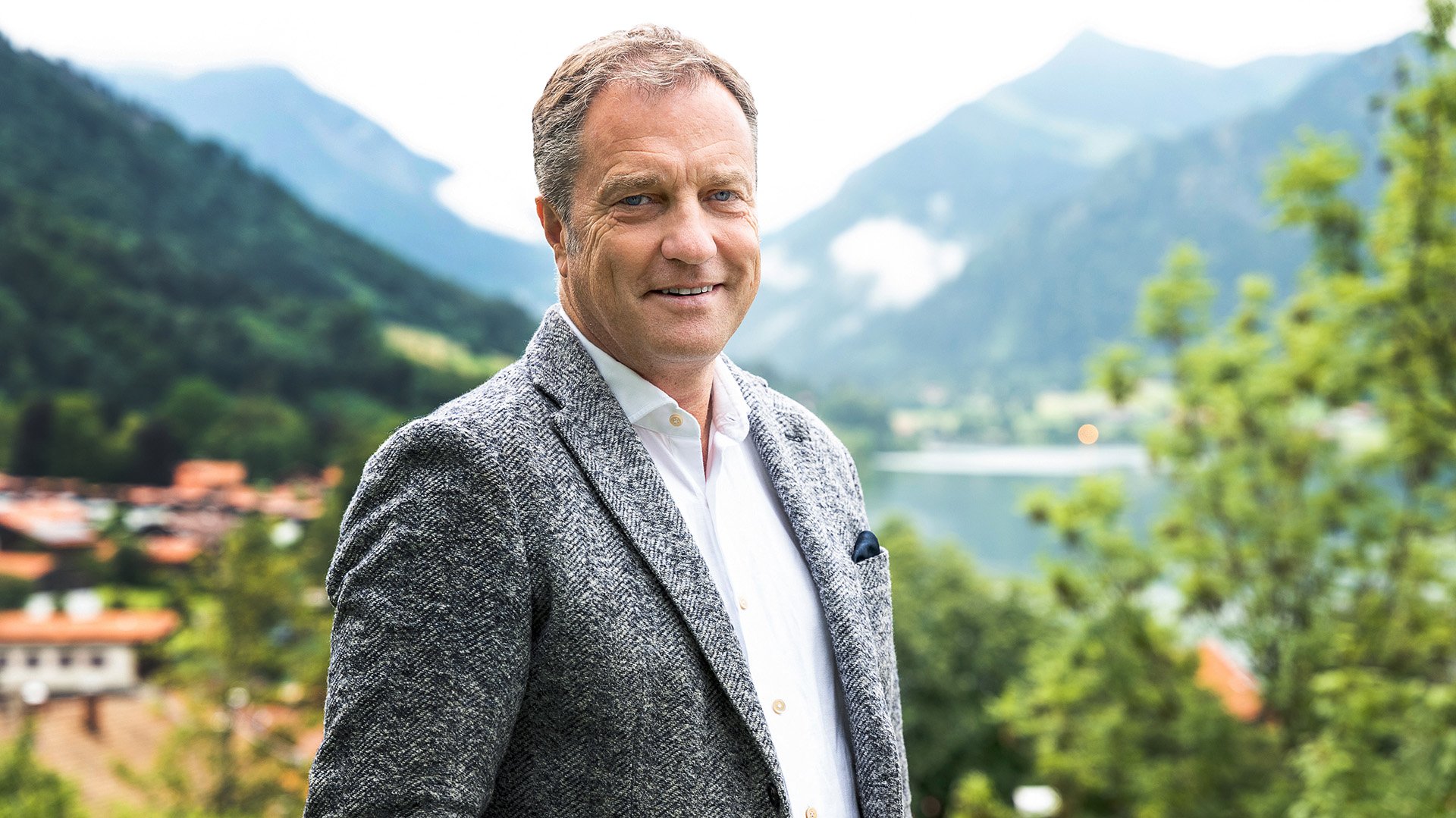 Porträtaufnahme einer Person in einem hellen Hemd und grauem Sakko, die im Freien vor einer alpinen Landschaft mit See, bewaldeten Hängen und Bergen steht; im Hintergrund sind zudem Dächer von Gebäuden und grüne Vegetation zu sehen.