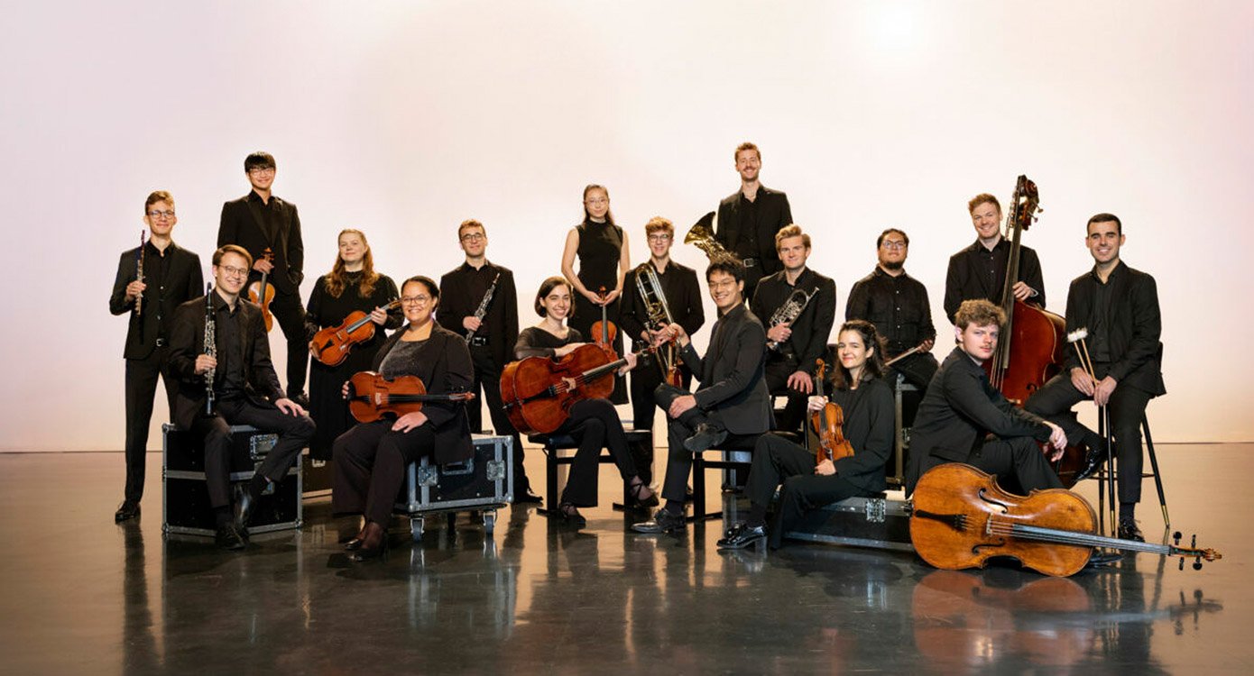 Das Orchester, dunkel gekleidet, mit Instrumenten posierend auf Bühne