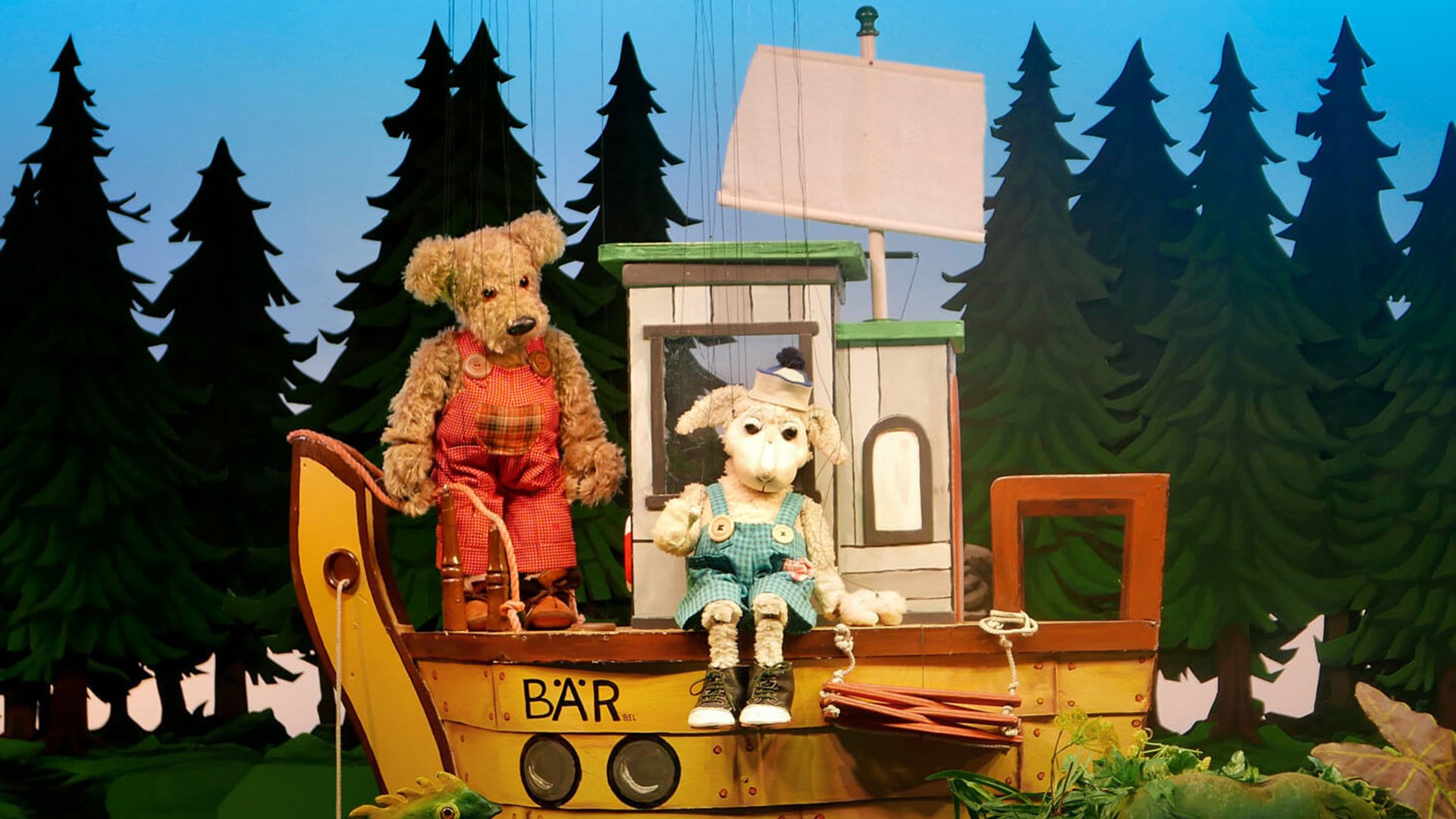 Puppenszene mit einem kleinen Bären in roter Latzhose und einem Lämmchen in türkisfarbener Latzhose, die auf einem gelben Boot mit der Aufschrift ‚BÄR‘ sitzen; im Hintergrund steht ein dichter Wald aus gezeichneten Tannenbäumen