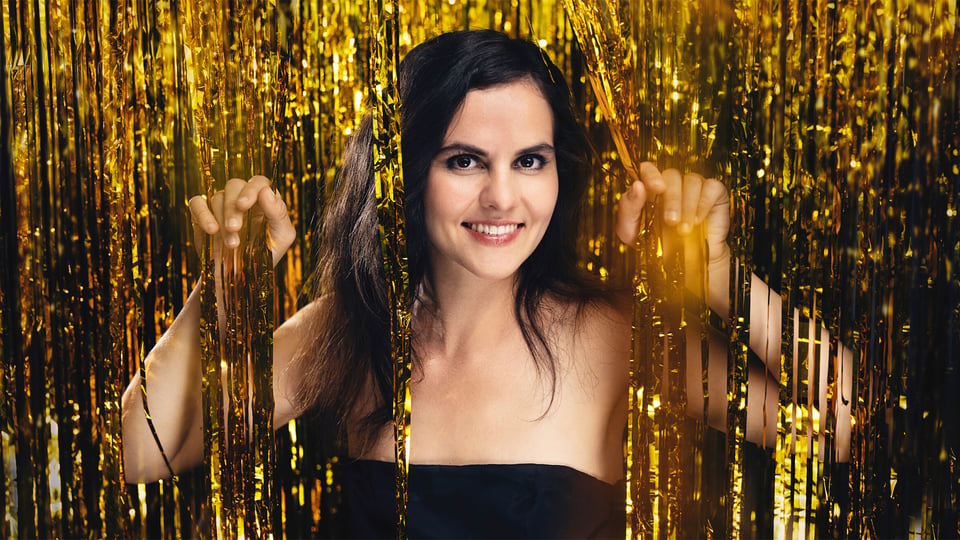 Eventmotiv zur „Festlichen Silvestergala“: Porträt einer Person vor einem goldfarbenen Lamettavorhang. Die Person steht frontal im Bild und hält die schimmernden Streifen mit beiden Händen leicht zur Seite. Dunkles, schulterfreies Bühnenoutfit, warme Beleuchtung mit goldenen Reflexionen. Glänzende, festliche Bildstimmung als Promomotiv für die Silvestergala im Herkulessaal der Residenz.