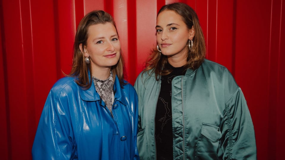 Zwei Damen, in blauer und grüner Jacke vor rotem Hintergrund