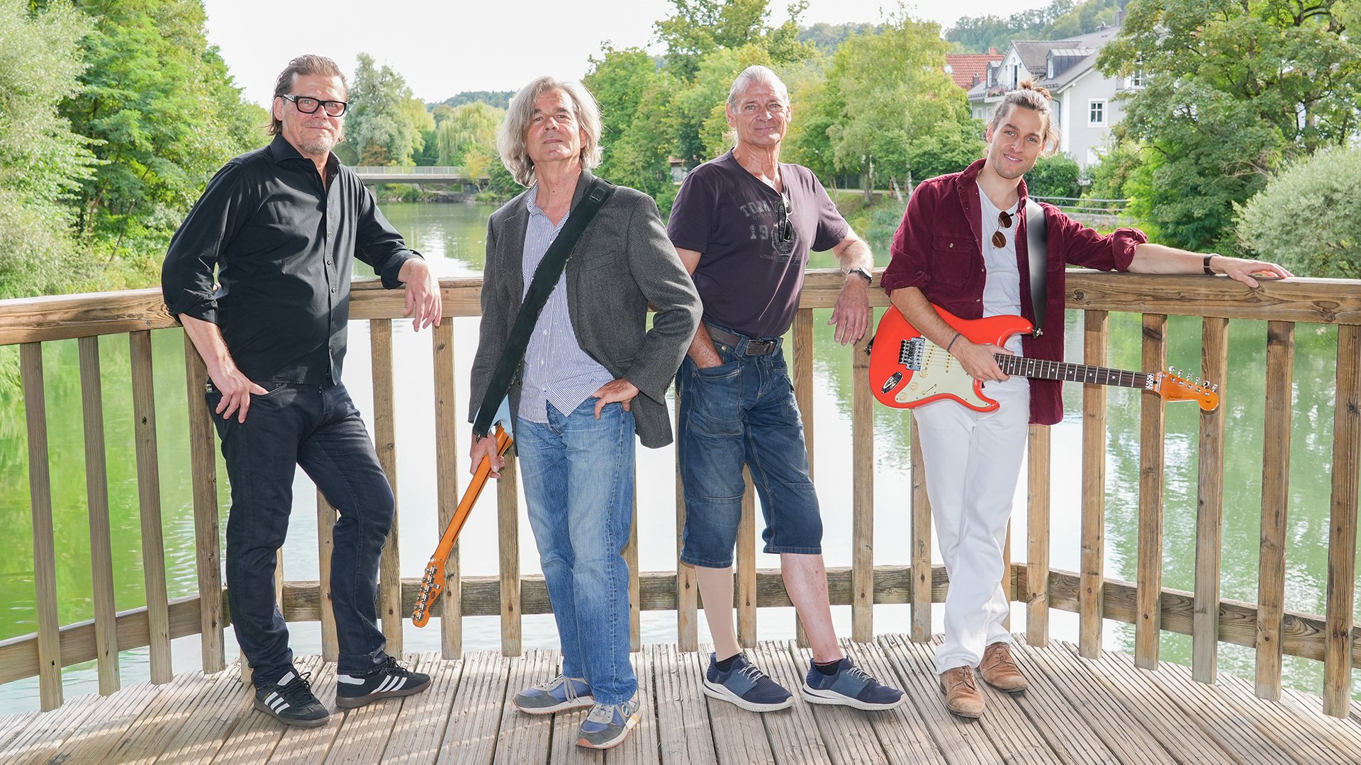 Bandfoto von Magic Buzz mit vier Musikern auf einer Holzplattform am Fluss. Zwei Personen halten E-Gitarren, im Hintergrund sind Bäume und Wohnhäuser zu sehen.