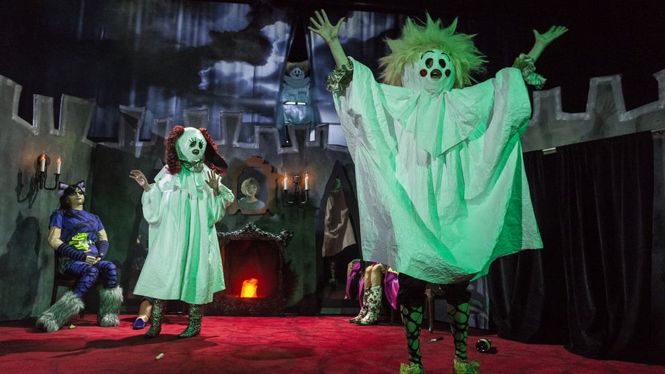 Halloweenartig inszenierte Theaterszene mit mehreren Darstellenden in Geisterkostümen vor einer gruseligen Kulisse mit Schlossmauern, Kerzenleuchtern und Kamin. Eine Figur mit wirrem Haar und ausgebreiteten Armen steht im Vordergrund, während weitere kostümierte Figuren auf der Bühne verteilt sind.