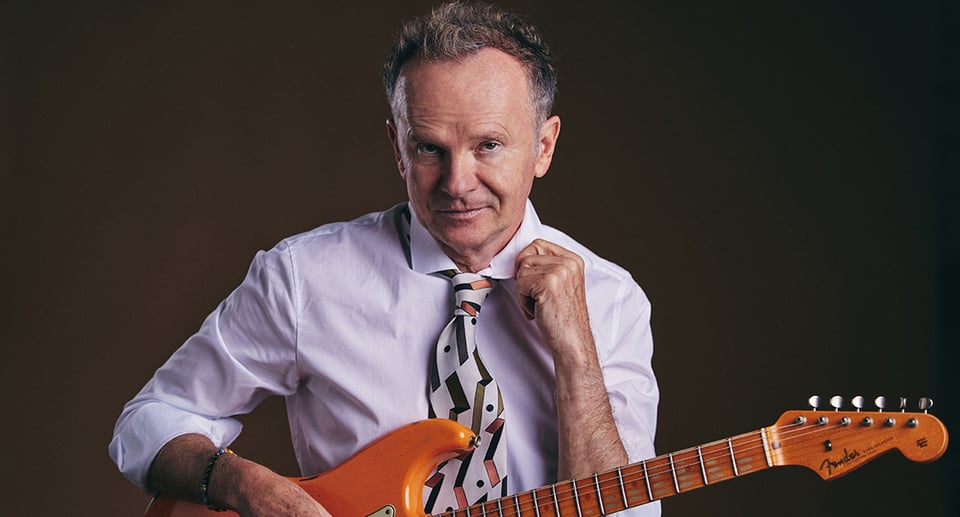 Bild des Künstlers mit weißer Krawatte und weißem Hemd und einer orangefarbenen E-Gitarre