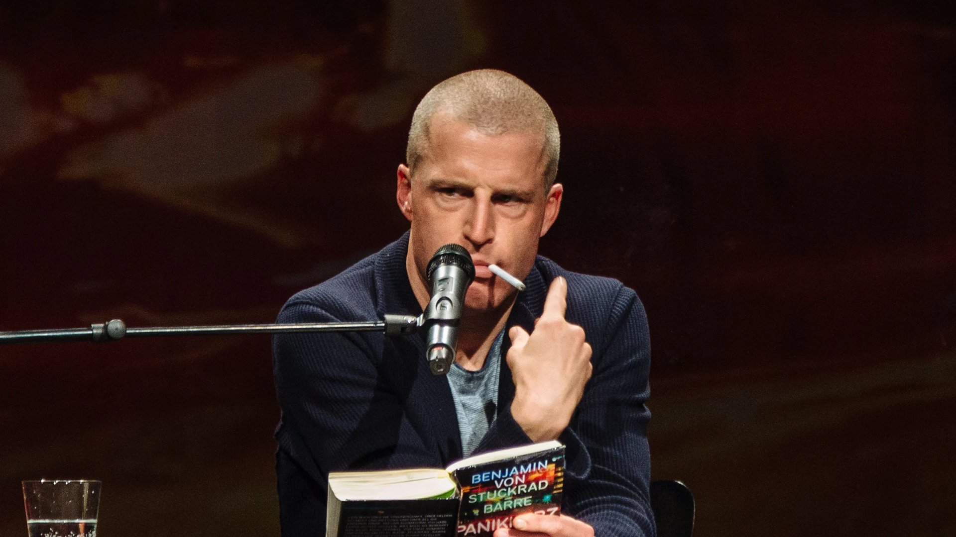 Benjamin Sturckrad-Barre liest mit einem Mikrofon aus seinem Buch vor und hat eine Zigarette im Mund