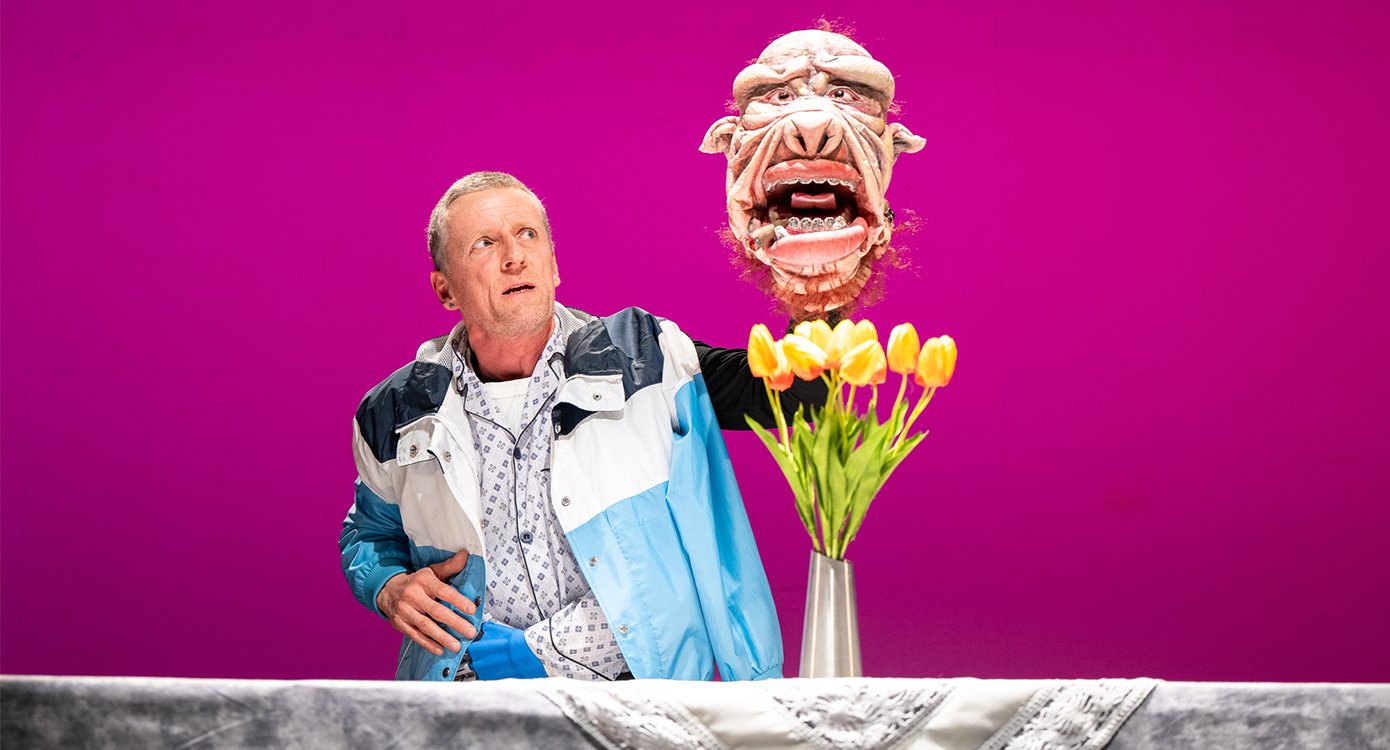 Foto des Künstlers mit einer Maske in der Hand und einer Vase mit gelben Tulpen vor pinkfarbenem Hintergrund.
