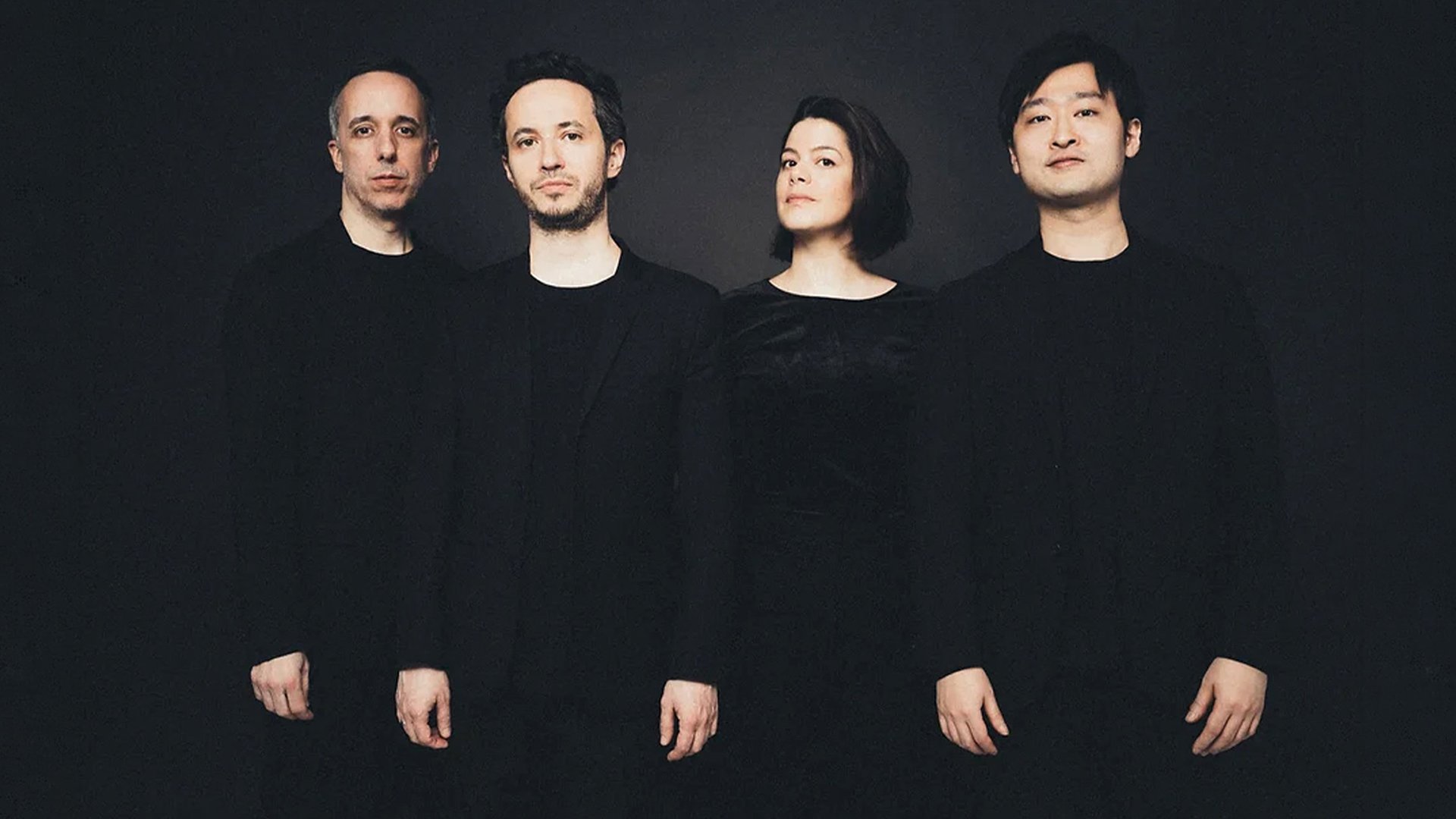 Quatuor Ébène mit vier Personen in dunkler Kleidung vor schwarzem Hintergrund, nebeneinander stehend in einer Studioaufnahme ohne sichtbare Instrumente.