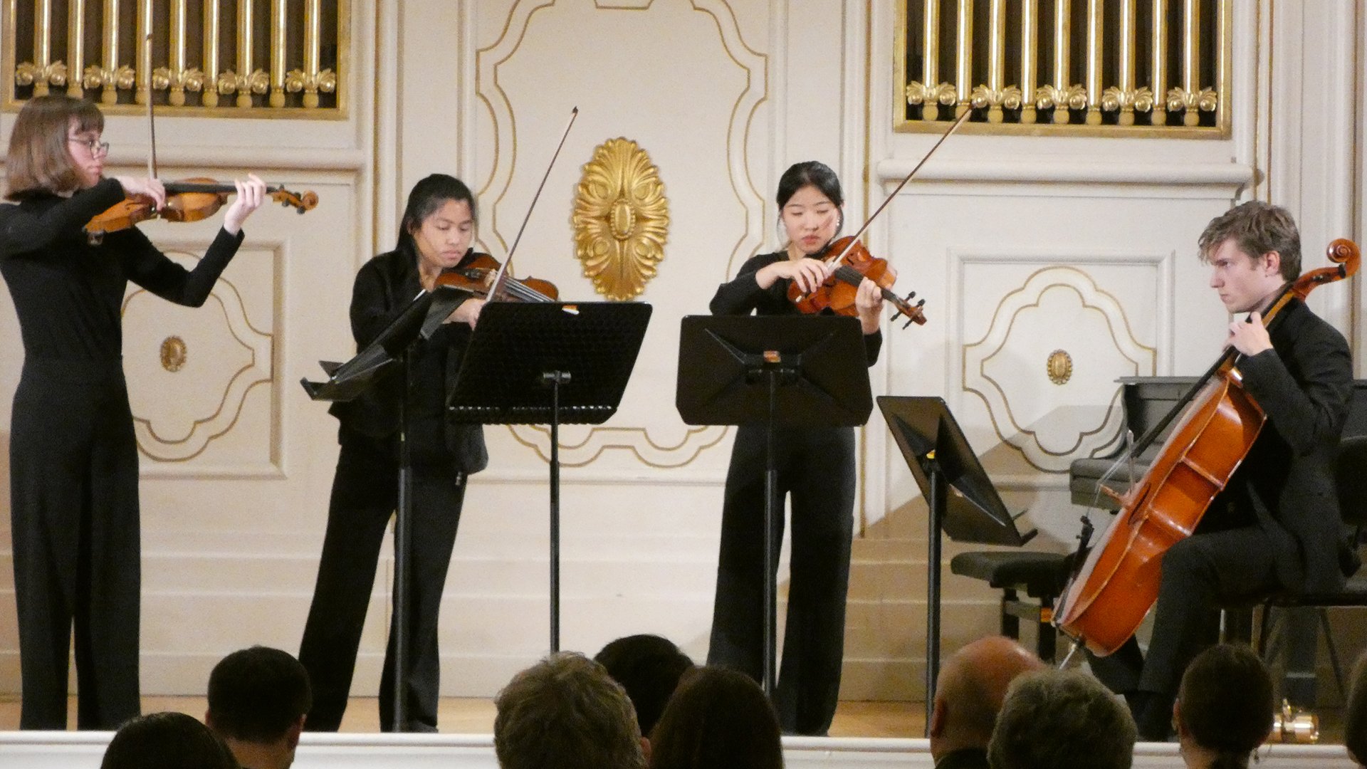 Vier Musiker in schwarzer Konzertkleidung spielen auf einer hellen Bühne mit weiß-goldenem Saalinterieur. Violinen, Viola und Cello sowie die warme Beleuchtung verleihen dem Konzert eine elegante, klassische Atmosphäre.