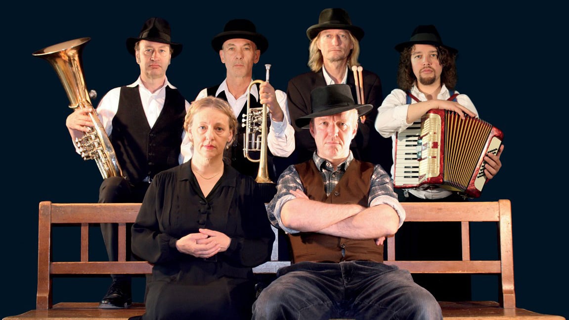Sechs Personen sind vor dunklem Hintergrund in einer inszenierten Gruppenaufnahme angeordnet: Zwei sitzen vorne auf einer Holzbank, vier stehen dahinter. Mehrere halten Musikinstrumente wie Tuba, Trompete, Schlagzeugstöcke und Akkordeon; alle tragen dunkle Kleidung mit Hüten.