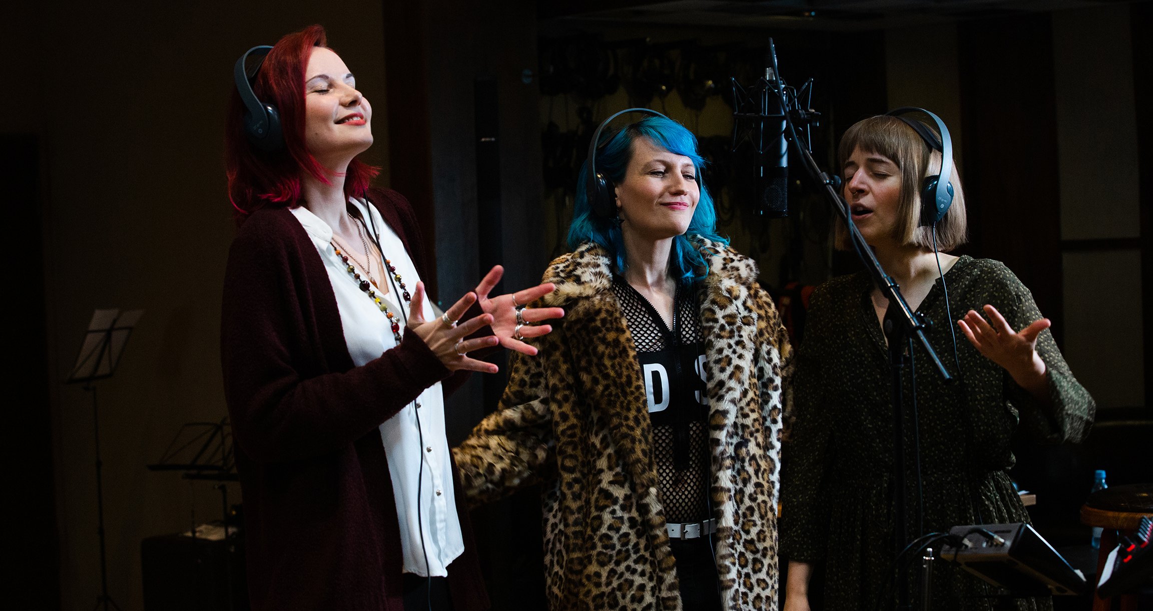 Foto von drei Mädchen, die in einem Aufnahmestudio singen. Ziemlich modernes Outfit, eine von ihnen mit blauem Haar.