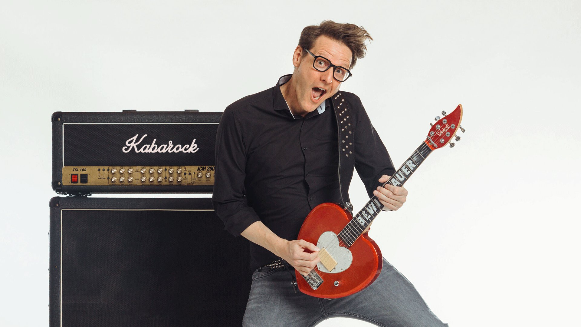 Bewie Bauer mit Brille auf spielt eine rote E Gitarre vor einem Verstärker mit der Aufschrift Kabarock und posiert in dynamischer Haltung vor weißem Hintergrund