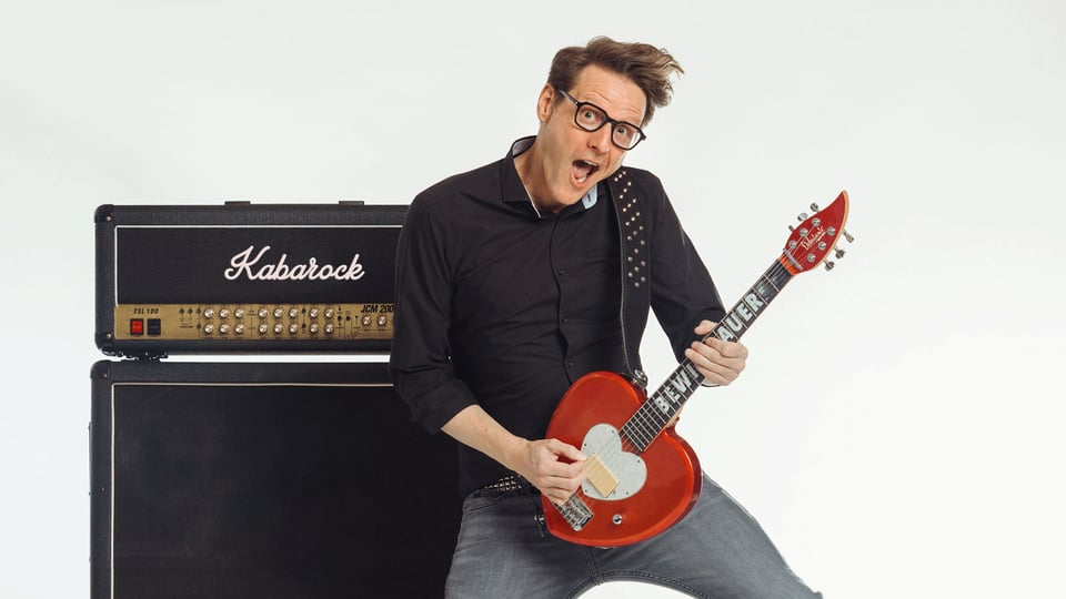 Bewie Bauer mit Brille auf spielt eine rote E Gitarre vor einem Verstärker mit der Aufschrift Kabarock und posiert in dynamischer Haltung vor weißem Hintergrund