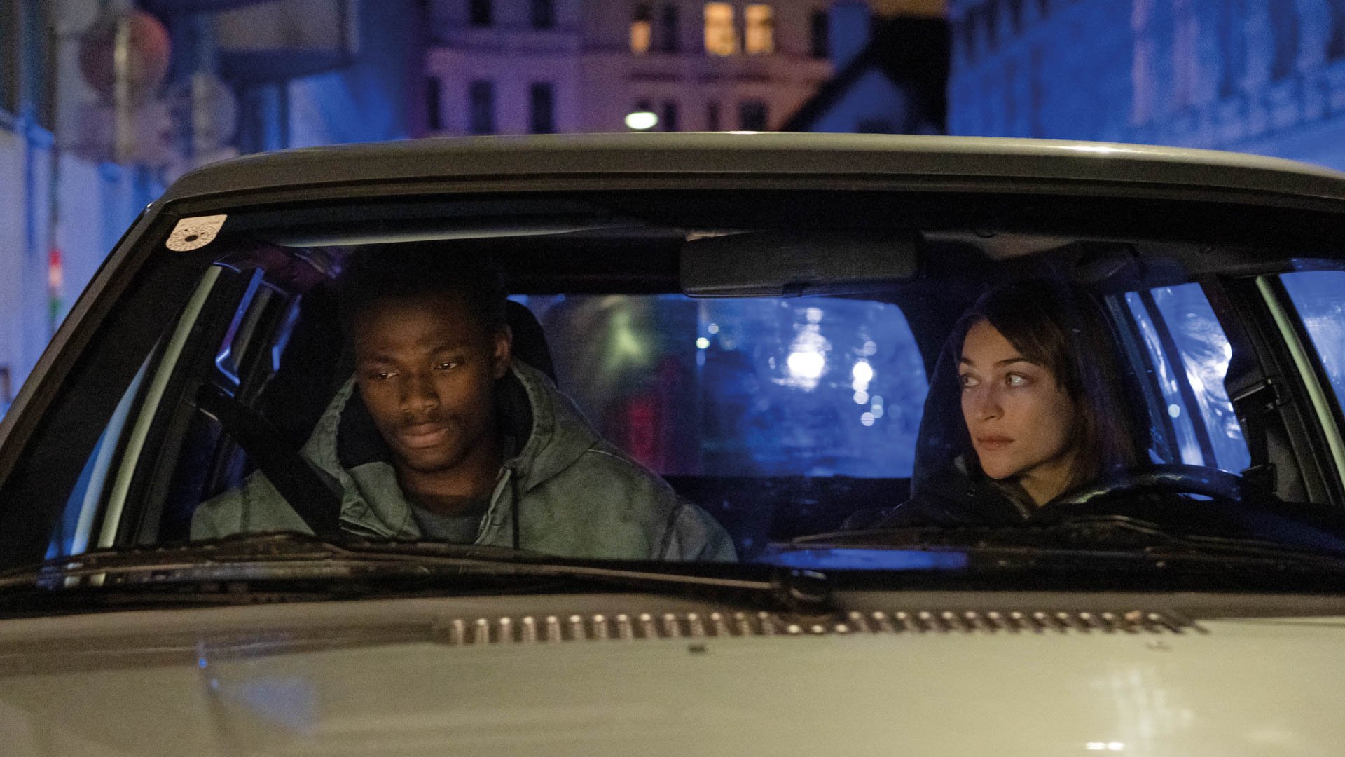 Zwei Personen sitzen nachts in einem Auto, beleuchtet von blauem Licht, und blicken nach vorne, im Hintergrund eine Straße mit Lichtern.