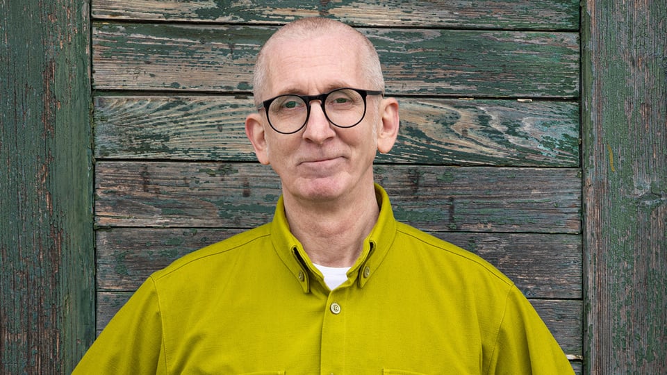 Der Kabarettist in einem grün-gelben Hemd vor einer rustikalen Holzvertäfelung