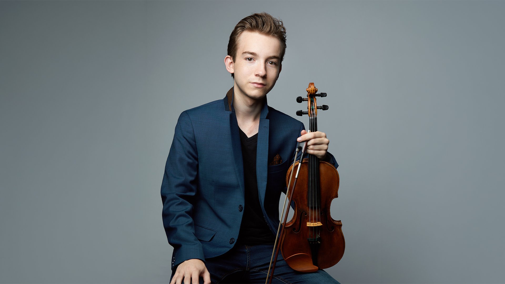 Porträtmotiv mit Violinisten und Violine: Eine sitzende Person hält eine Violine senkrecht vor dem Körper und blickt frontal in die Kamera. Dunkler Blazer über schwarzem Oberteil, neutral grauer Studiohintergrund mit weicher Ausleuchtung. Klarer, reduzierter Bildaufbau; Promofoto für ein klassisches Orchester‑ und Violinkonzert mit den Münchner Symphonikern.