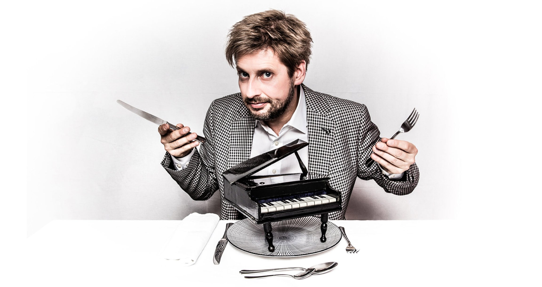 Foto des Künstlers Daniel Helferich, wie er an einem Tisch sitzt, Besteck in den Händen hält und auf dem Teller ein kleines Klavier liegt – alles vor weißem Hintergrund.